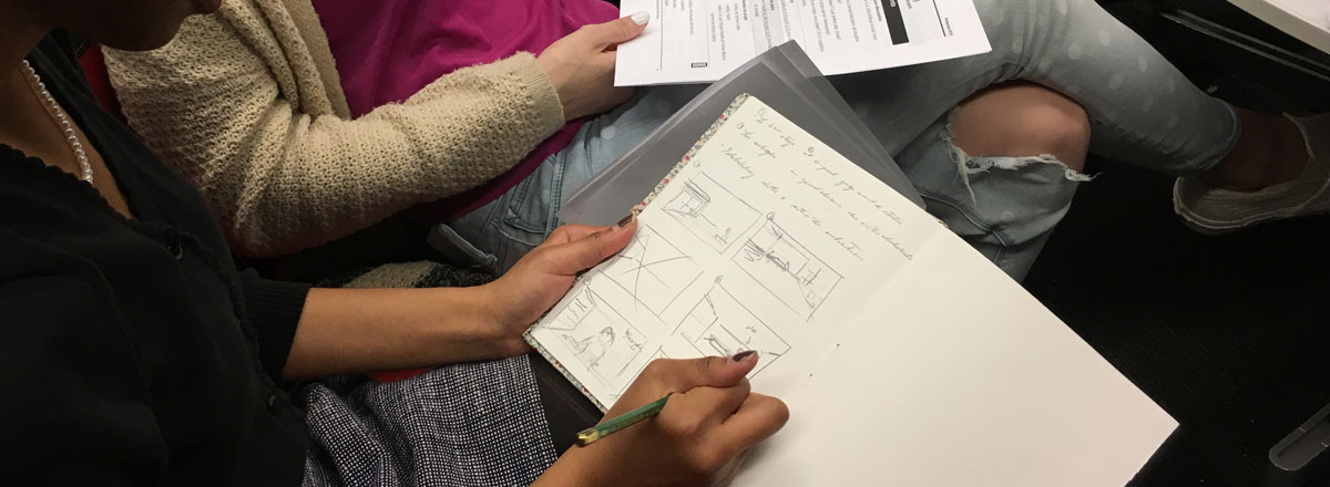 An instructor sketches wireframes in a notebook while students look over her shoulder