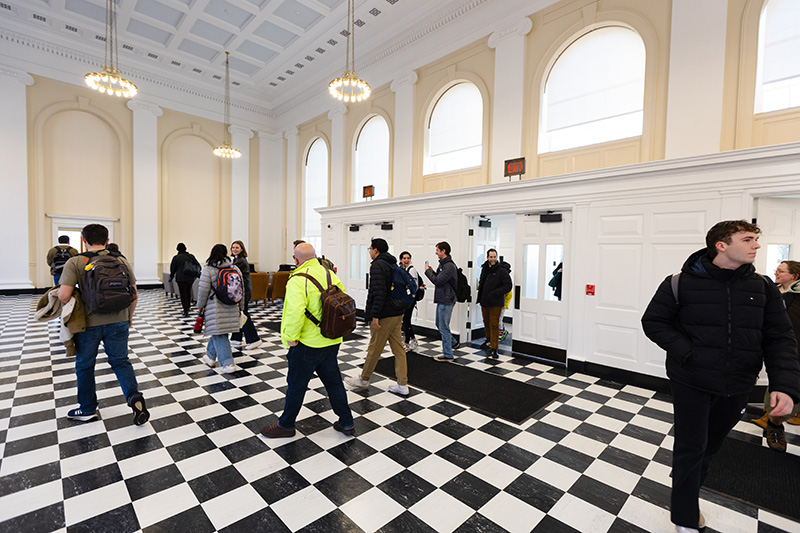 A group of people walking out of a vestibule and pouring into a large room. The room has light fixtures on long chains hanging from very high ceilings, and black and white checkered floors.