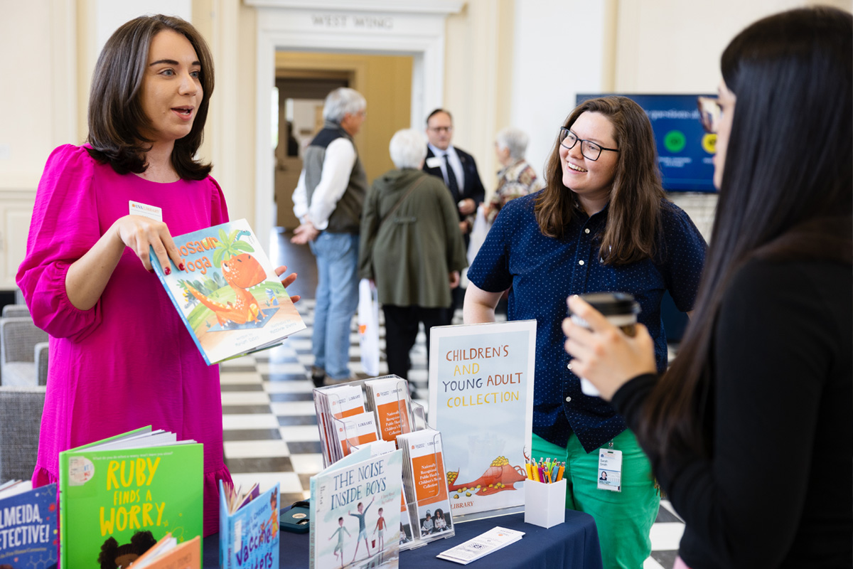 Three individuals engaging in conversation beside a table showcasing children's books, including titles like “Rory the Rabbit” and “The Kids’ Book of Questions.” One person is handing a book to another while gesturing. The event is held in a bright, spacious hall with arched doorways.