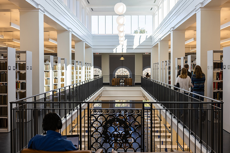 People, some seated, some walking, in a brightly lit library with shelving, multiple windows, and an aperture in the floor surrounded by a railing.