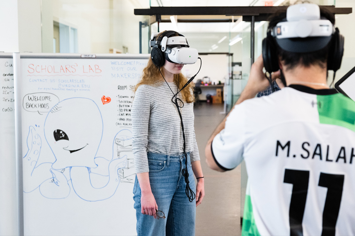 Person wearing a virtual reality headset standing near a whiteboard labeled Scholars’ Lab while another person observes.