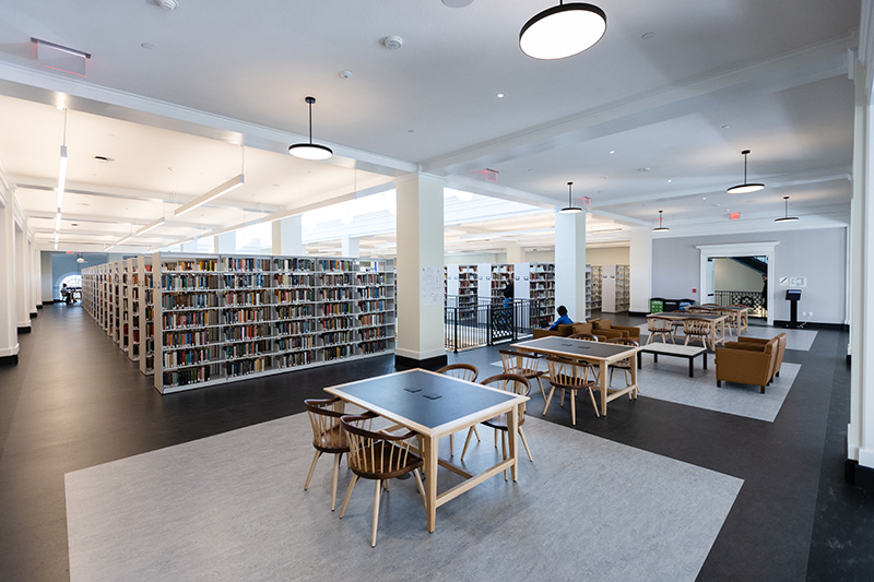 An open seating space in a library with wooden tables and chairs, as well as armchairs and low coffee-table style tables.