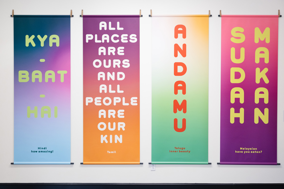 Four colorful banners hanging on a white wall, each displaying a positive phrase in different languages: “Kya Baat Hai” in Hindi (meaning “how amazing!”), “All places are ours and all people are our kin,” a Tamil phrase written in English, “Andamu” in Telugu (meaning “inner beauty”), and “Sudan Makan” in Malaysian, meaning “have you eaten?”