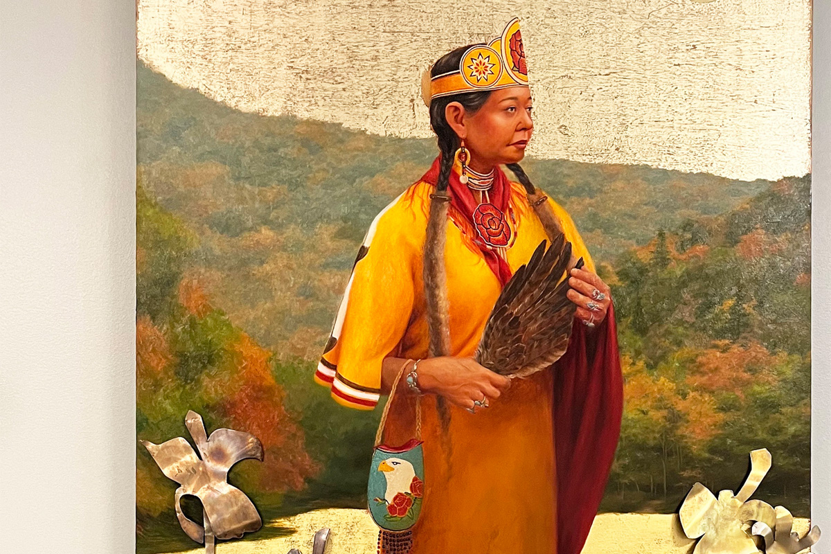 Portrait of a person in traditional Native American attire, holding a feather and standing before a backdrop of autumn trees.