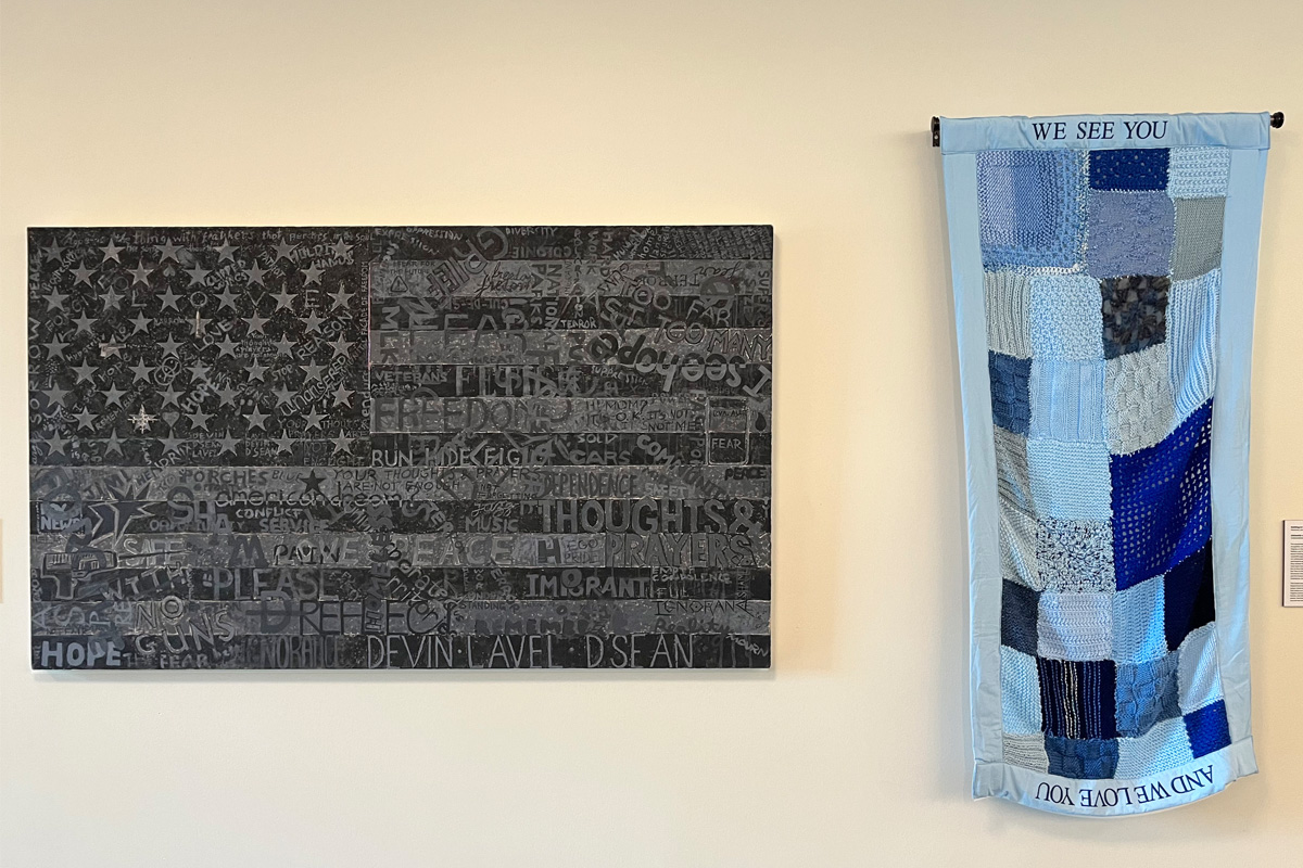 Two art pieces displayed on a wall. On the left, an American-flag themed painting features dark tones with star patterns and text in various fonts and sizes. On the right, a vertical banner with a blue and grey patchwork design includes the text “WE SEE YOU” at the top and “AND WE LOVE YOU” at the bottom.