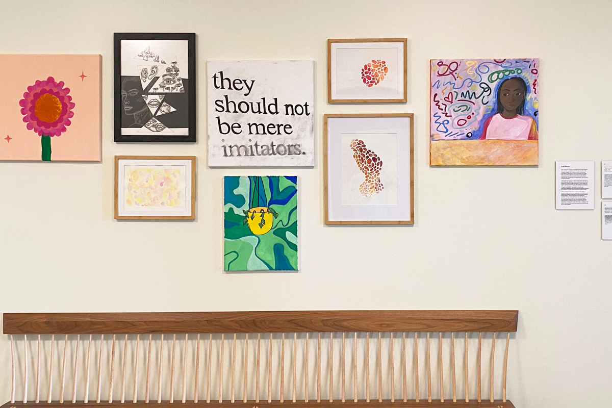 Gallery wall featuring a diverse array of eight artworks including floral patterns, textual art with the phrase ‘they should not be mere imitators’, abstract pieces, and a portrait, displayed above a wooden bench.