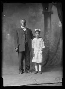 A man in a suit and vest stands with his hand on a young girl's shoulder. The girl wears a plaid dress and a hair bow. 