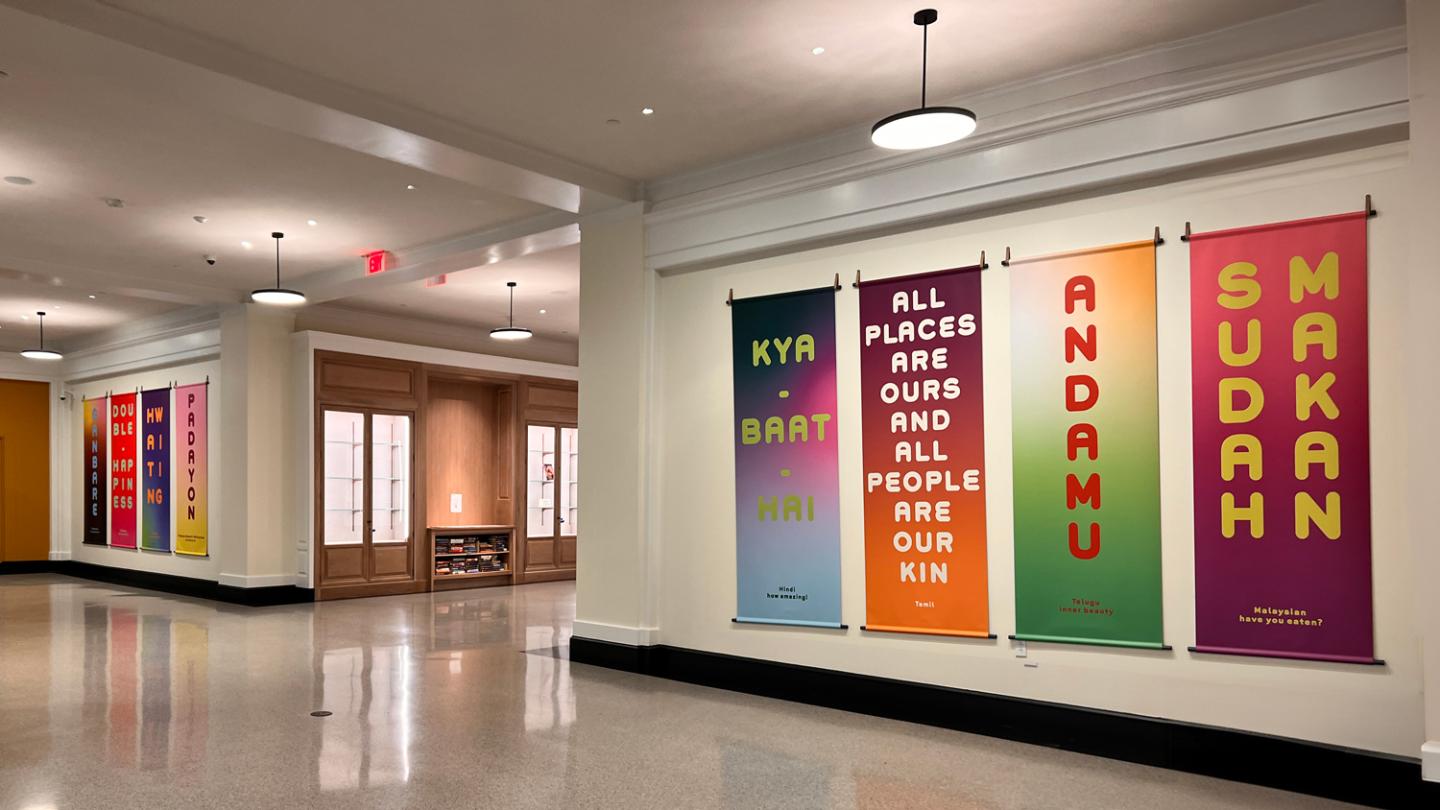 Eight colorful vertical panels are on display in an open hallway. They read phrases in various languages, each translating to a positive or uplifting message.