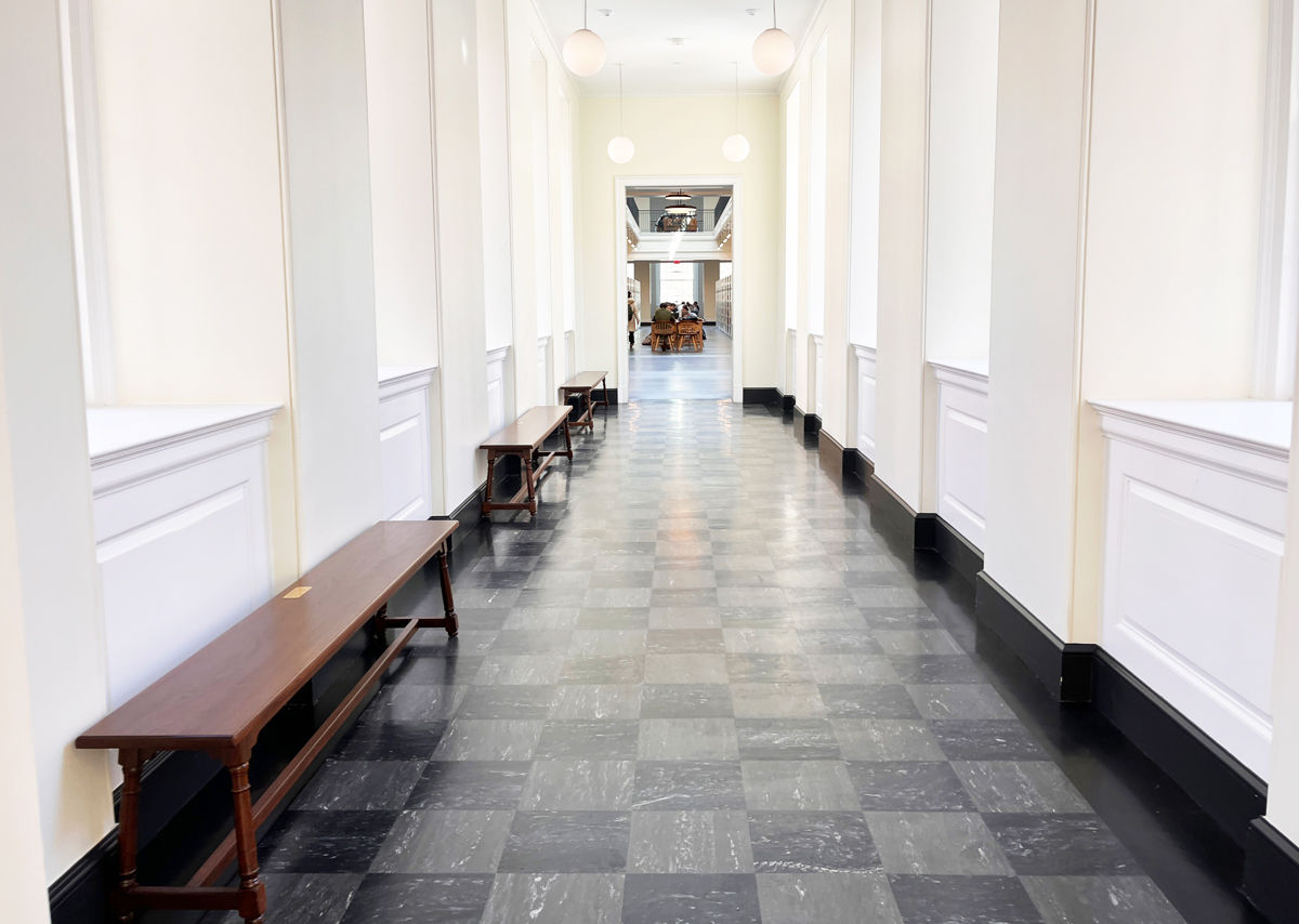 A long, elegant hallway with large windows on the left, casting natural light onto a floor of checkered tiles. Wooden benches line the walls, and spherical ceiling lights hang above. The hallway leads to a bright open area with tables and library shelving.