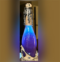 A decorative glass bottle filled with a glowing blue liquid. The bottle is topped with a cork covered in black wax, which has dripped down the sides. A small skull adorns the top of the cork, adding a spooky effect. The base of the bottle is wrapped in a bone-like material, enhancing the eerie, gothic aesthetic. The background is blurred, drawing focus to the illuminated bottle.