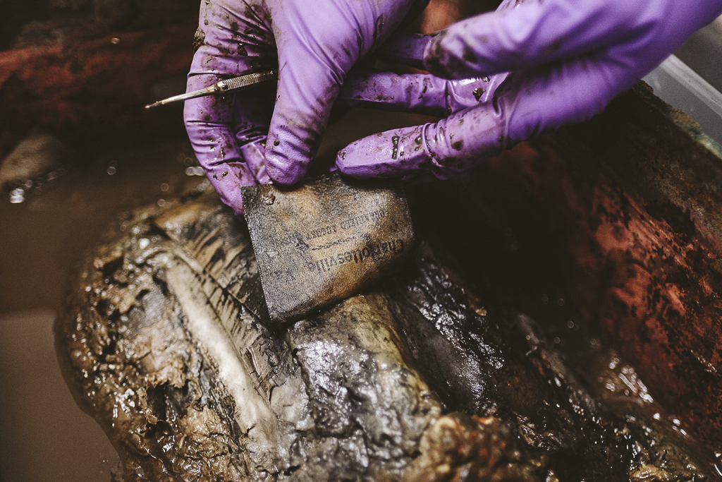 Gloved hands holding tweezers carefully peel back a ribbon from the contents of the open time capsule