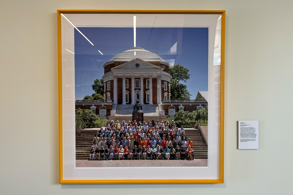 A framed photograph on a wall depicting a large group of people seated on steps in front of the iconic Rotunda, a large building of red brick with a white dome and white columns, at the University of Virginia.