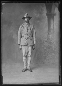 A young man wearing military dress, a flat-brimmed hat, and glasses looks directly at the camera. 