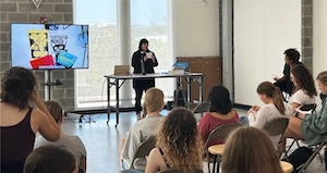 A person presenting to a group of seated attendees in a room with a large screen displaying a PowerPoint slide.