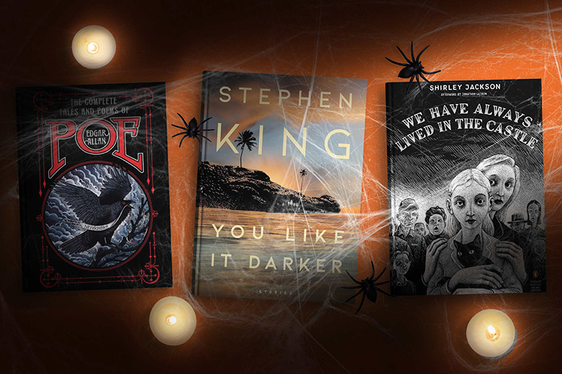 A spooky Halloween-themed display of three gothic literature books surrounded by candles, cobwebs, and black plastic spiders on a dark surface. The books include "The Complete Tales and Poems of Edgar Allan Poe," featuring a raven illustration on the cover; "You Like It Darker" by Stephen King, with an eerie beach scene; and "We Have Always Lived in the Castle" by Shirley Jackson, displaying a haunting illustration of a girl holding a cat with a ghostly expression. The setup evokes a dark, atmospheric vibe perfect for Halloween.