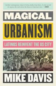 The cover of a book titled "Magical Urbanism: Latinos Reinvent the US City" by Mike Davis. The title is in bold, all-caps font, with "Magical" in white text on a dark gray background and "Urbanism" in black text on a yellow background. Below, there is a pink strip with the subtitle "Latinos Reinvent the US City" in white text. The bottom section features a black-and-white photo of a large crowd of people holding protest signs. At the bottom, "Mike Davis" is written in bold white text on a dark gray background.