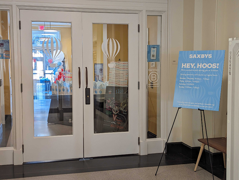 Glass doors are closed in front of a cafe with the Saxbys logo on the doors. A blue sign beside the door lists business hours. 