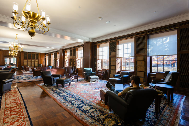 A student lounges in a comfortable chair in in an ornate, old-fashioned room with Persian rugs and a fireplace.