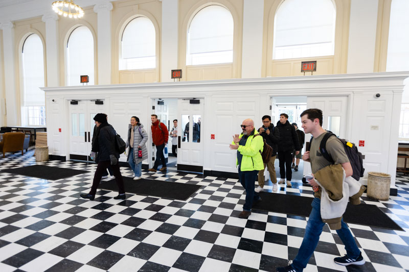 Twelve people enter a large room through the front doors. A man in a green coat applauds.