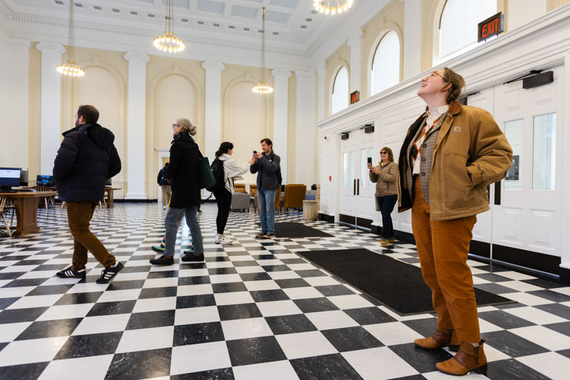 A woman stands in the lobby of the library gazing up in happiness. Beind her, people take pictures and mingle. 