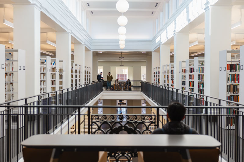 Students gather around an apeture railing on the fifth floor of the library, overlooking the fourth floor below. Above them, natural light flows in through clerestory windows.