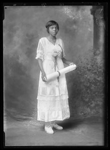 A young woman stands, holding a large white scroll with a bow around it. She is wearing a delicate white dress and fresh flowers, looking directly at the camera.