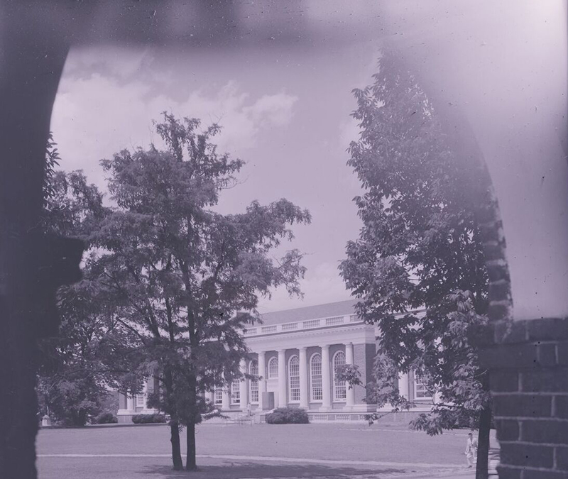 A black-and-white photograph of a large neoclassical-style building with tall columns and large arched windows, partially obscured by tall trees in the foreground. The scene is viewed through an arched opening, adding a frame around the image. The photograph has a slightly eerie, foggy effect, giving it a vintage and mysterious atmosphere.